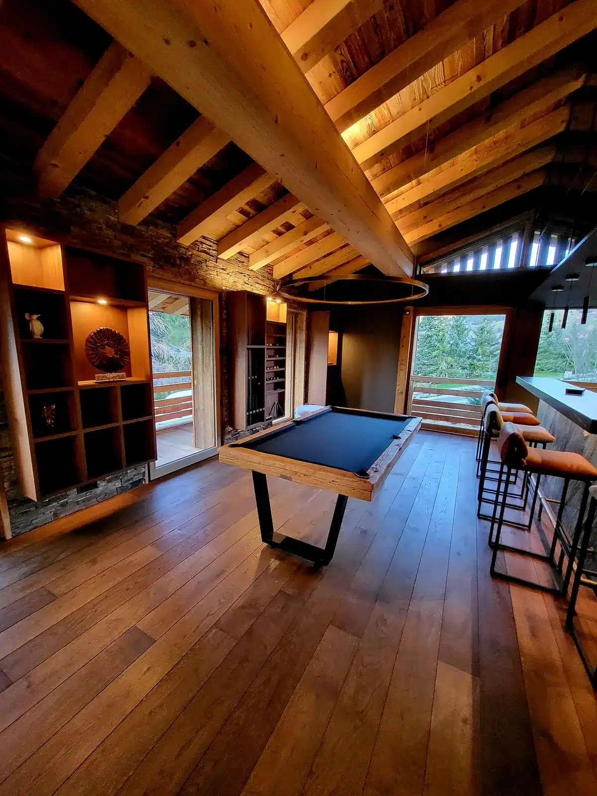 Salle de jeux rustique-chic sous un toit cathédrale avec poutres apparentes, billard moderne, bar et vues extérieures boisées.