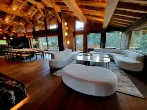 Grand salon de chalet de luxe avec poutres apparentes en bois, canapé incurvé blanc moelleux, table basse moderne et salle à manger attenante.