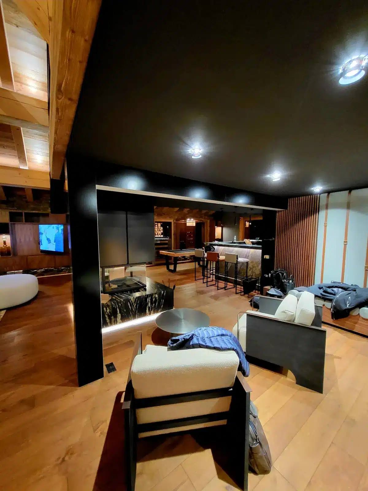 Intérieur luxueux style chalet avec plafond noir, plancher en bois chaud, foyer moderne en marbre, espace bar et table de billard visibles au fond.
