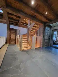 Escalier bois massif et verre dans un hall de chalet. Plafond à poutres, murs rustiques, et grand carrelage gris ardoise au sol.