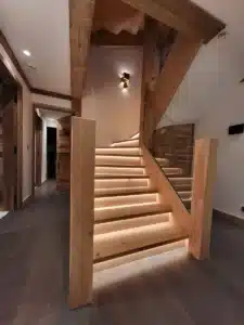 Magnifique escalier en bois rustique éclairé par LED sous les marches. Poutres en chêne massif et rambarde en verre dans un hall moderne.