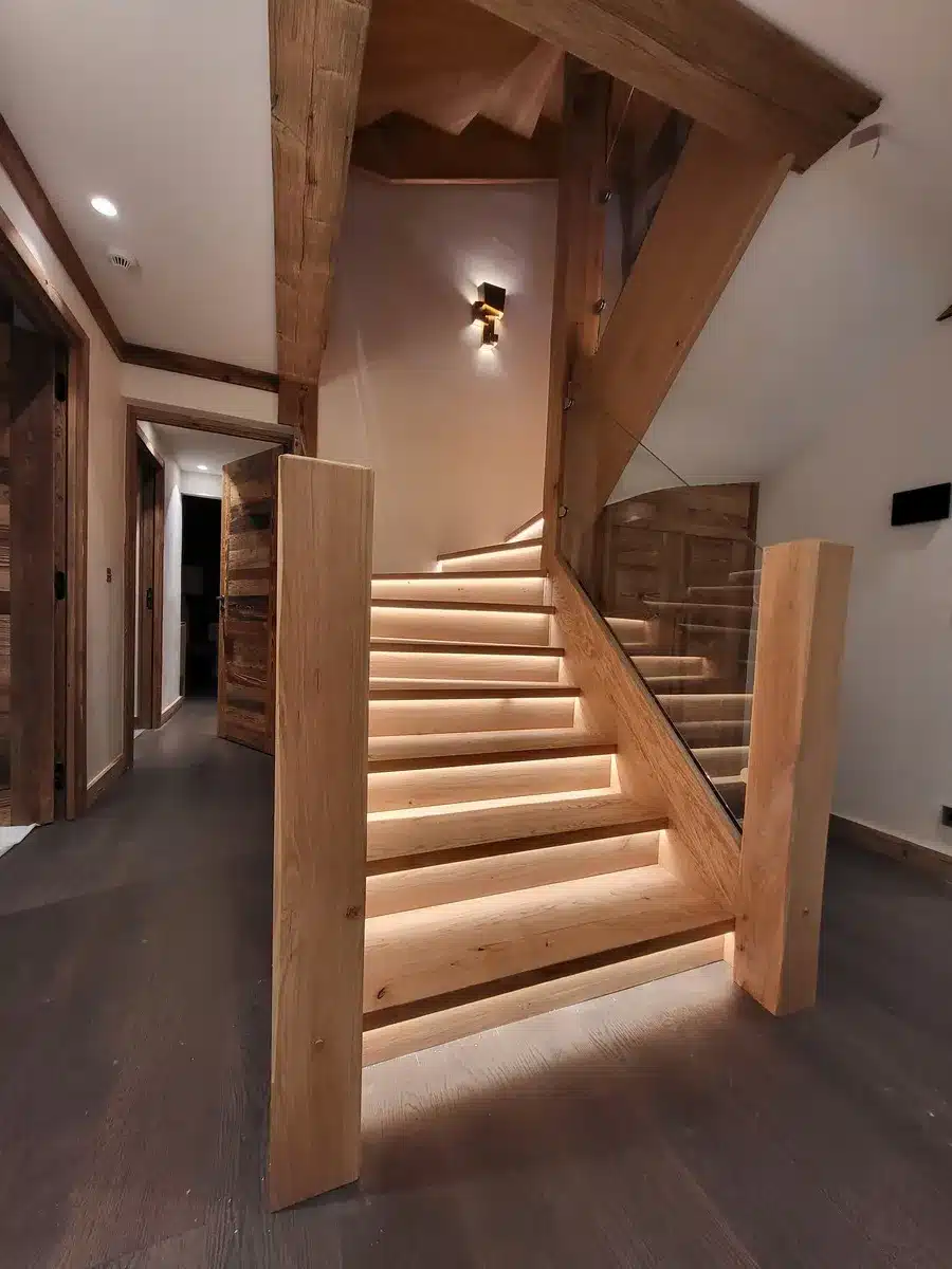 Magnifique escalier en bois rustique éclairé par LED sous les marches. Poutres en chêne massif et rambarde en verre dans un hall moderne.