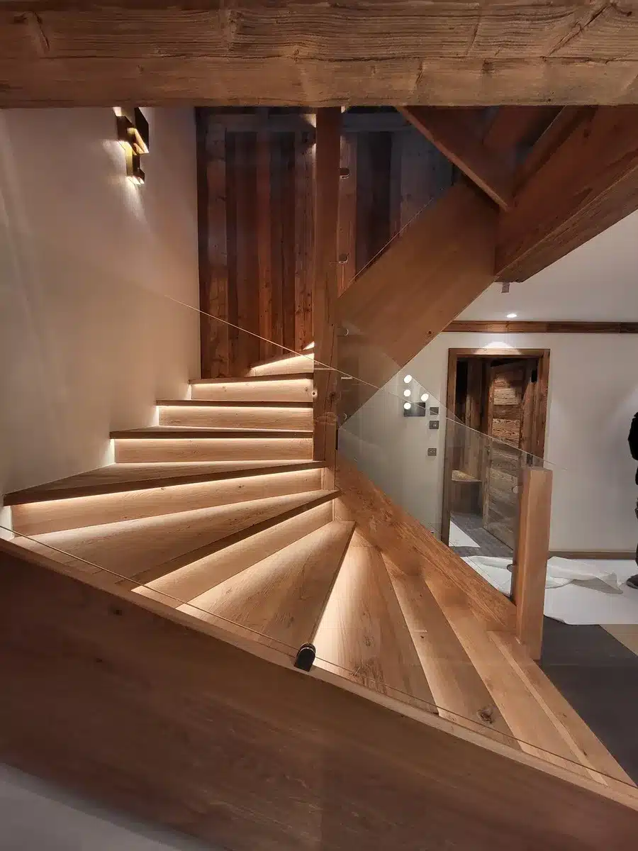 Escalier en bois tournant, éclairage LED intégré sous les marches. Garde-corps en verre et poutres rustiques de style chalet.