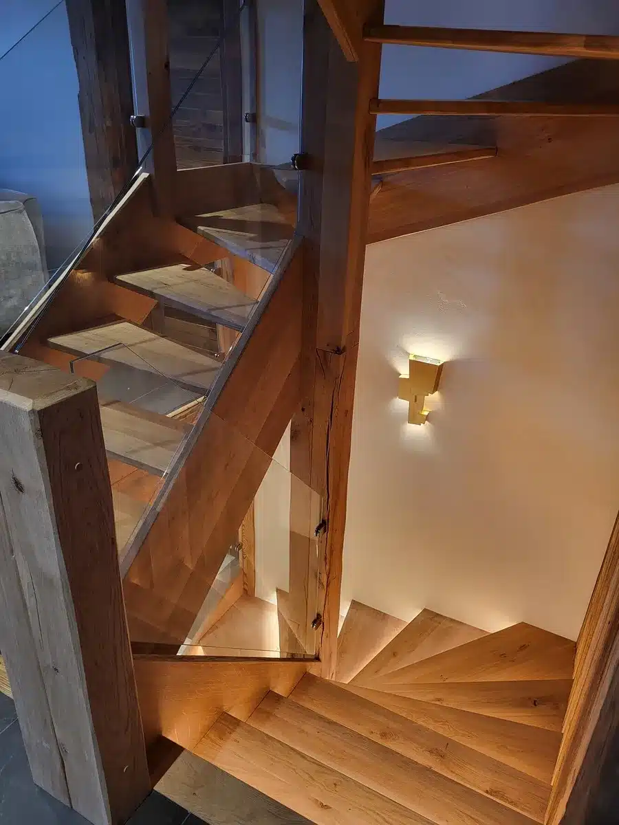 Escalier bois massif et verre. Design contemporain, marches tournantes éclairées par une applique murale croisée chaleureuse.