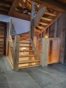 Escalier ouvert en bois brut et verre dans un intérieur chalet. Le design rustique moderne met en valeur les marches flottantes et les poutres apparentes.