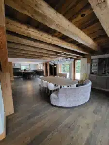 Intérieur de chalet rustique-chic: poutres anciennes et plancher foncé. Salle à manger ouverte avec banquette courbe, table en bois clair et lustre bulles.