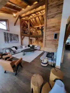 Luxueux salon de chalet : poutres en bois, cheminée en pierre, canapé gris moderne et fauteuils en cuir caramel sur plancher foncé.